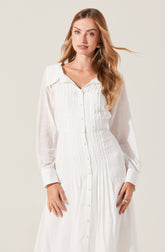 A woman is wearing a long, white dress with a button-down front and a collar. The dress features vertical pleats running down the front and long, semi-sheer sleeves with wide cuffs. The fabric appears lightweight and slightly textured.